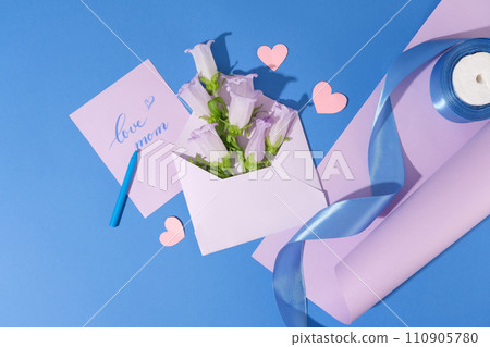 Envelope of several fresh flowers displayed with a card with text love mom. A roll of purple paper with blue ribbon roll placed on arranged with a crayon and paper hearts. Concept for special event 110905780