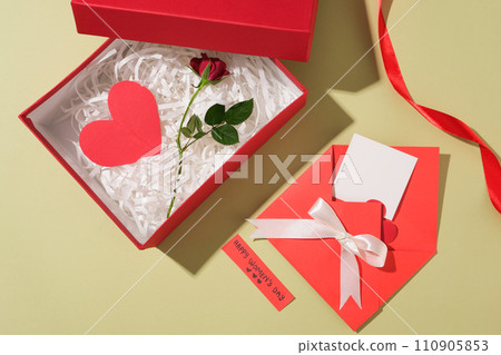 A gift box in red color containing a lot of kraft paper with a red paper heart and a branch of rose placed on. Red envelope with a ribbon bow displayed with a small paper of wishes A gift box in red color containing a lot of kraft paper with a red paper heart and a branch of rose placed on. Red envelope with a ribbon bow displayed with a small paper of wishes 110905853