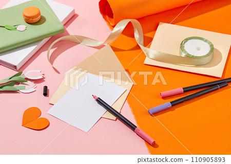 Several color markers displayed with a blank piece of paper and envelopes. A stack of notebooks featured with a roll of ribbon. Paper cut in tulip flowers and heart shaped. Women Day concept 110905893