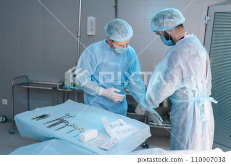 Side view of assistant male helping surgeon putting on latex gloves before operation. Team of surgeons in operating room with light modern interior preparing for surgery. Concept of emergency. Side view of assistant male helping surgeon putting on latex gloves before operation. Team of surgeons in operating room with light modern interior preparing for surgery. Concept of emergency. 110907038