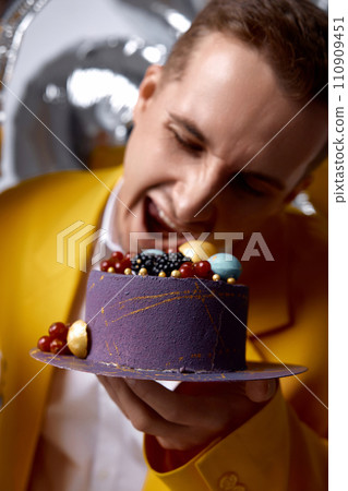birthday man in yellow jacket bites cake on helium balloons background. birthday man in yellow jacket bites cake on helium balloons background. 110909451