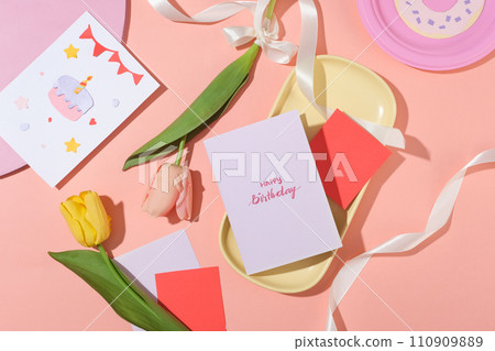 Top view of the words happy birthday on a white card. Cute cards, ribbons and tulips on a pink background. Creative space with birthday party theme. Top view of the words happy birthday on a white card. Cute cards, ribbons and tulips on a pink background. Creative space with birthday party theme. 110909889