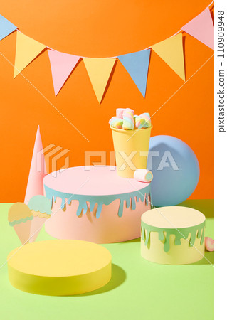 Front view of two birthday cakes made from colored paper, a yellow blank podium, a marshmallow cup, origami cream and a blue balloon. A decorative triangular flag string on an orange background. 110909948