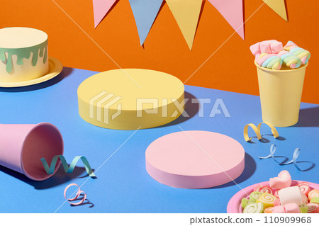 Two cylindrical platforms are displayed on a blue surface with paper cups, ribbons and marshmallows. The orange background is decorated with a triangular flag string. 110909968