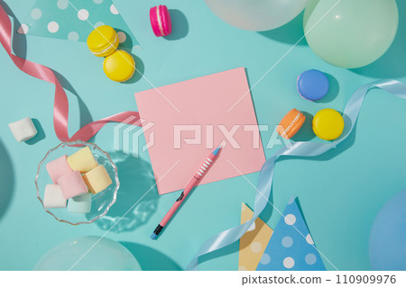 A pencil sits next to a pink card. Marshmallows, ribbons, balloons and macarons are decorated around the blue background. Free space to design birthday wishes. 110909976