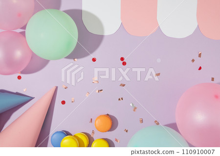 On a pastel purple background, balloons, birthday hats, macarons and confetti are decorated. Empty space for good birthday wishes design. Top view. 110910007