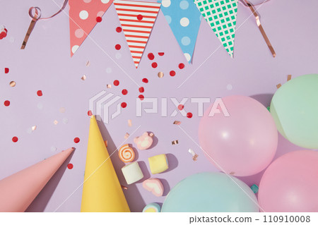 Flat lay of a string of triangular flags, birthday hats, balloons, marshmallows and confetti on a pastel purple background. Brilliant birthday space. 110910008
