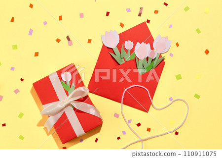 View from above of colorful confetti on pastel yellow background. A gift box and a red envelope with tulips cut from colored paper are displayed. 110911075