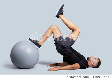 Concentrated muscular young caucasian guy with beard, make exercises with fit ball for legs, enjoy workout Concentrated muscular young caucasian guy with beard, make exercises with fit ball for legs, enjoy workout 110911453