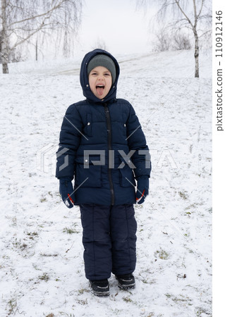 Funny little kid in warm overalls sticking out his tongue and grimacing in winter park. Funny little kid in warm overalls sticking out his tongue and grimacing in winter park. 110912146