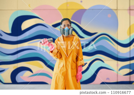 Female graffiti artist in respirator mask standing near the wall with her paintings looking to camera 110916677