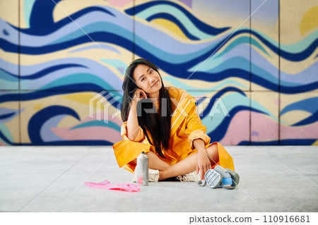 Female street artist rest sitting on the ground near the wall with her colorful paintings 110916681