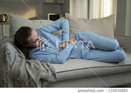 Sick, unhappy young brunette suffers from abdominal pain in everyday life, lies at home on the couch holding her stomach, experiences abdominal pain and discomfort, suffers from menstrual cramps 110916892