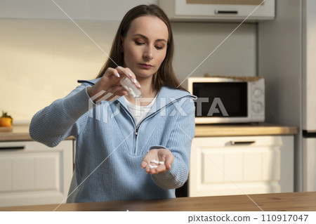 Woman pours pills into the palm of her hand while taking medicine sitting at the kitchen table Woman pours pills into the palm of her hand while taking medicine sitting at the kitchen table 110917047