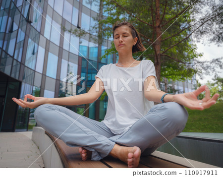 De-stress, unwind idea. peaceful female freelance employee practicing yoga exercises while sitting in bunch under glass skyscrapers after hard working day in office, holding hands in mudra De-stress, unwind idea. peaceful female freelance employee practicing yoga exercises while sitting in bunch under glass skyscrapers after hard working day in office, holding hands in mudra 110917911
