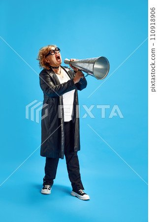 Adult elderly woman dressed in leather trench coat shouting loudly to vintage megaphone to attract people's attention Adult elderly woman dressed in leather trench coat shouting loudly to vintage megaphone to attract people's attention 110918906