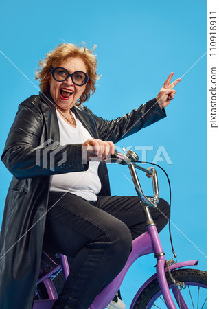 Feel like child. Elderly woman expressing fun and joy while riding in motion on children's bicycle against blue background. 110918911
