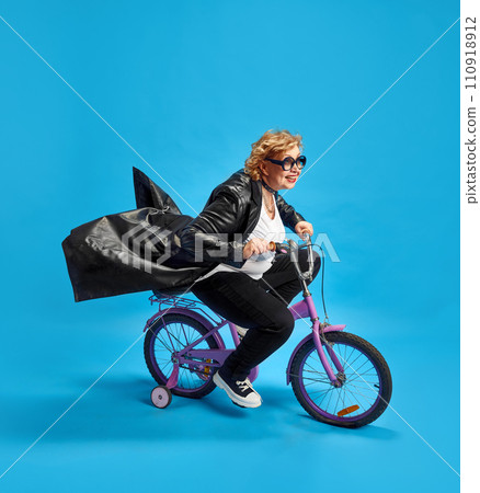 Elderly woman exuding coolness and joy while riding in motion on children's purple bicycle against blue background. Elderly woman exuding coolness and joy while riding in motion on children's purple bicycle against blue background. 110918912