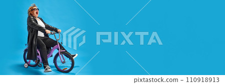 Elderly woman exuding coolness and joy on purple bicycle dressed leather jacket, sunglasses against blue background with negative space. 110918913