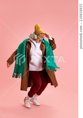 Charming, excited elderly woman in fashion outfit dancing while listening energetic music against pink background. 110918915