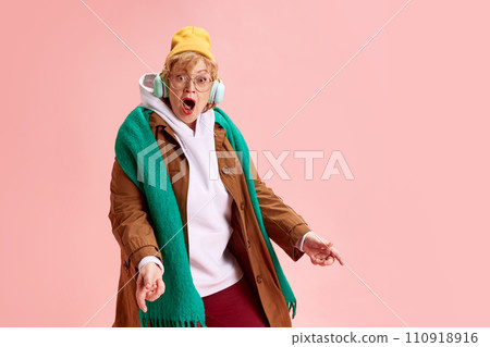 Portrait of senior lady dressed fashion outfit and glasses listening music in headphones against pink background. Model looks surprised. 110918916