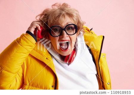 Emotional senior woman listening music in headphones and singing wearing yellow puffer jacket, white shirt against pink background. 110918933
