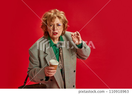Picky mature woman, looks as boss, looking at camera with cup of coffee and asking something to fix against red background Picky mature woman, looks as boss, looking at camera with cup of coffee and asking something to fix against red background 110918963