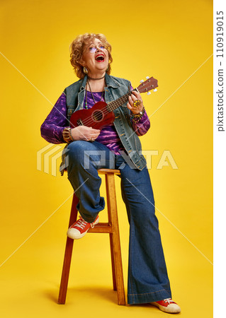 Joyous senior woman energetic playing ukulele seat on stool dressed casual denim and red shoes against yellow background. 110919015
