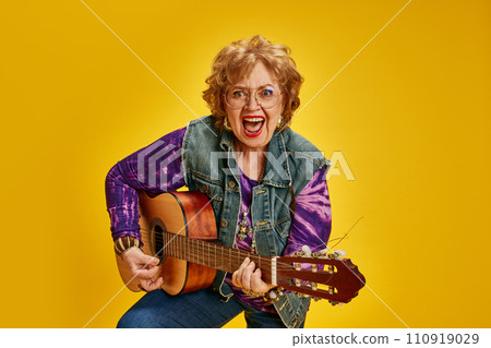 Portrait of elderly lady playing guitar with excited and joyful expression dressed denim attire, tie-dye fashion T-shirt. Portrait of elderly lady playing guitar with excited and joyful expression dressed denim attire, tie-dye fashion T-shirt. 110919029