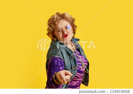 Senior woman puckering lips in round glasses dressed denim jacket, purple T-shirt with gold jewelry against yellow background. 110919088