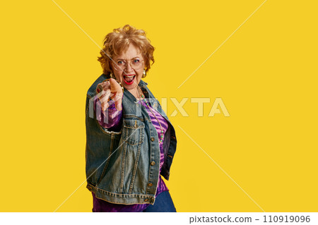 Joyful senior woman pointing at camera dressed in denim jacket, purple blouse against yellow background. 110919096