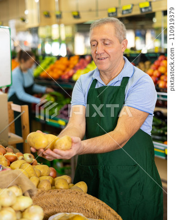Aged salesman putting potatoes on food stall in greengrocery Aged salesman putting potatoes on food stall in greengrocery 110919379
