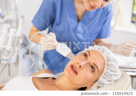 Young female client experiencing facial skin peeling using ultrasonic scrubber 110919423