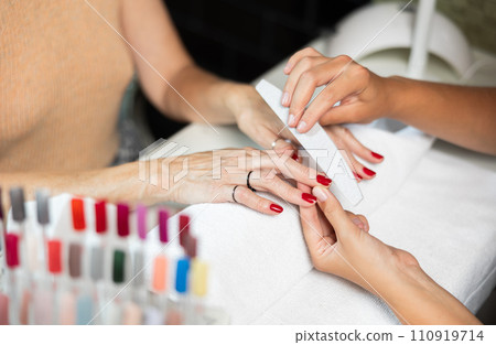 Hands of young girl manicurist perform trimming saws nails to elderly lady client. 110919714