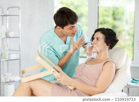 Male beautician giving consultation to woman patient with mirror in aesthetic clinic Male beautician giving consultation to woman patient with mirror in aesthetic clinic 110919733