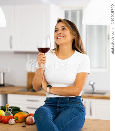 Smiling young Hispanic woman drinking wine sitting on table in kitchen 110919740