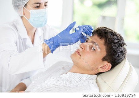 Professional cosmetologist hands in gloves performing lips enhancement treatment to male client, making injection of lip fillers Professional cosmetologist hands in gloves performing lips enhancement treatment to male client, making injection of lip fillers 110919754