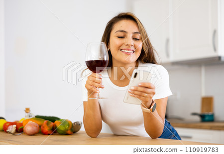 Housewife with glass of red wine in kitchen reading messages on mobile phone screen Housewife with glass of red wine in kitchen reading messages on mobile phone screen 110919783