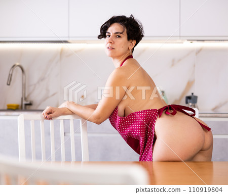 Seductive shorthaired woman dressed only in apron standing near chair in kitchen 110919804