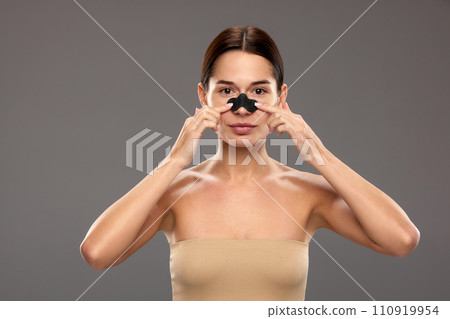 Young woman taking care after skin, applying nose mask for black spots, pores cleansing against studio background 110919954