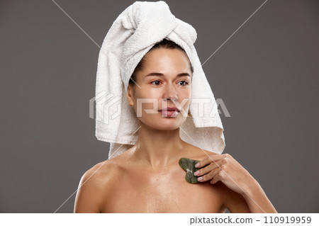 Beautiful young woman in towel on head, after shower doing body massage with gua sha against studio background 110919959