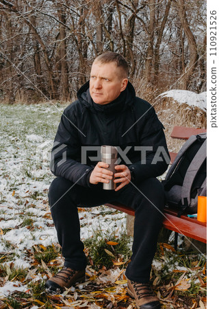 Winter Serenity: 40-Year-Old Man Enjoying Tea on Snow-Covered Bench in Rural Park. Immerse yourself in the tranquil beauty of winter as a 40-year-old man finds solace on a snowy bench in a rural park. 110921526