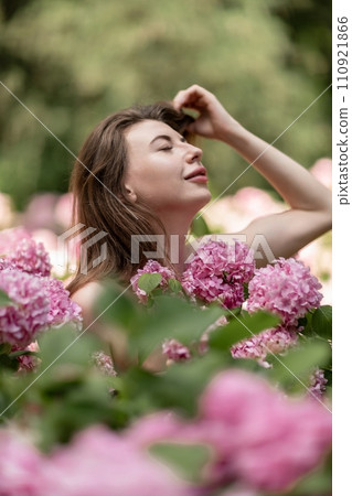 Hydrangeas Happy woman in pink dress amid hydrangeas. Large pink hydrangea caps surround woman. Sunny outdoor setting. Showcasing happy woman amid hydrangea bloom. 110921866