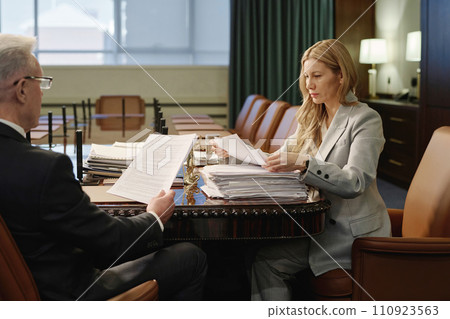 Female lawyer and her senior colleague sitting at wooden desk in boardroom and reading documents 110923563