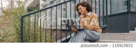 Pretty female entrepreneur working on laptop sitting outside on modern building background 110923652