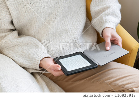 A man in a cozy sweater reads an ebook Kindle on a digital tablet, sitting comfortably. The setting is indoors and creates a warm and comfortable atmosphere. 110923912