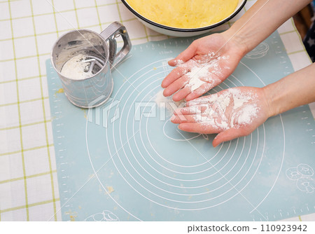 The image shows hands covered in flour over a silicone baking mat. A sifter and mixing bowl are nearby. The image shows hands covered in flour over a silicone baking mat. A sifter and mixing bowl are nearby. 110923942