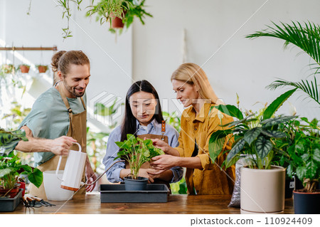 Busy working day at flower shop. Tree coworkers, asian woman, young man and middle age successful blond female owner together. Caucasian handsome male watering plant. 110924409