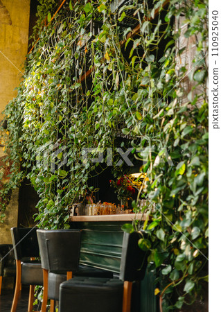 Interior of modern loft style restaurant. Bar counter covered in vivid greenery ivy, high wooden leather chairs. Cozy place design decorated with houseplants in eco cafe indoor. Interior of modern loft style restaurant. Bar counter covered in vivid greenery ivy, high wooden leather chairs. Cozy place design decorated with houseplants in eco cafe indoor. 110925040