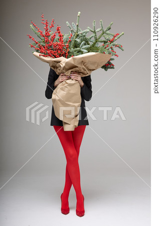 A long girl in red tights and high-heeled shoes holds a huge bouquet of fir branches and red berries wrapped in cardboard paper, she closes with a bouquet, photo studio with white background A long girl in red tights and high-heeled shoes holds a huge bouquet of fir branches and red berries wrapped in cardboard paper, she closes with a bouquet, photo studio with white background 110926290